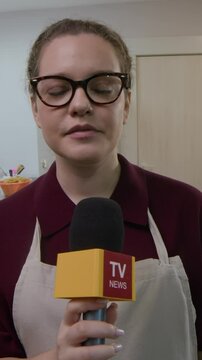 Vertical waist up of young female baker in beige apron and glasses speaking into microphone during interview inside clean kitchen neatly arranged with baking tools and trays