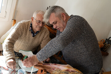 father and son in their fifties cutting iberian ham leg, family activity teaching how to cut it...