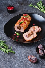 Ready-made pork tenderloin roll with figs and walnuts, wrapped in bacon, on a black ceramic plate on a dark concrete background. Recipes pork.