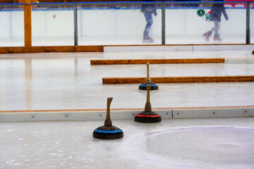 Three curling stones with wooden handles are positioned on an icy surface, while blurred figures...