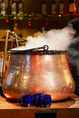 A steaming copper cauldron stands on a wooden table, surrounded by blue mugs, with a warm festive...
