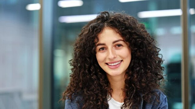 Cheerful businesswoman with curly hair takes off glasses and smiles at the camera in the office. Stylish woman in a blue jacket on a blurred office background.