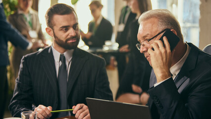 Senior businessman multitasking by discussing project on phone while collaborating with young man,...