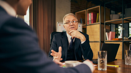 Focused businessman in his late 50s sitting at table in office, engaged in lively conversation with young male employee. Providing constructive feedback and offering help. Business, mentorship concept