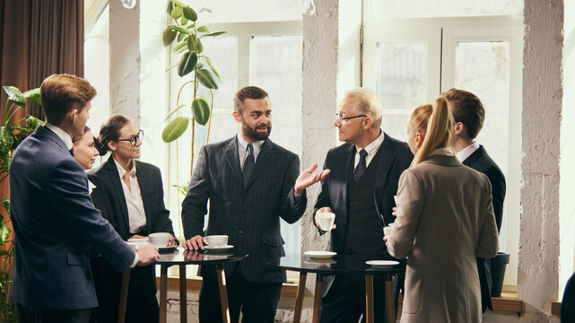 Group of young colleagues in formal wear talking with senior businessman in friendly conversation during coffee break. Exchange of ideas and team building. Concept of business, office, teamwork