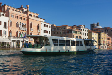 A white passenger boat sails on a canal in front of colorful historic buildings with arched windows and balconies, under a clear blue sky, creating a picturesque scene.