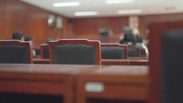 A classroom with a few people sitting in chairs. The chairs are black and the room is wooden