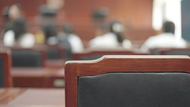 A wooden chair with a black seat is in a room with many other chairs. The room is filled with people, some of whom are sitting and others standing. The atmosphere of the room seems to be formal