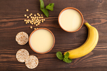 Organic non dairy banana and soy milk in clay cup on brown wooden, top view, close up.