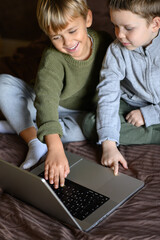 The photo shows two young boys sitting on their bed in their room, engaged in an online lesson with a teacher via a laptop. They are focused and interacting with the screen, while the cozy atmosphere 