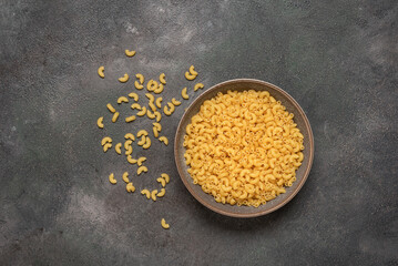 Raw elbow macaroni pasta in bowl on dark grunge background, top view.