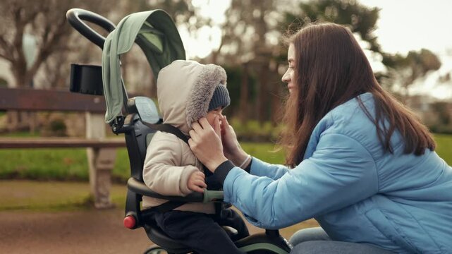 Little toddler son sits in a tricycle stroller in the park, mom approaches, fixes his clothes, hugs and kisses him. Happy motherhood and child care