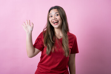 Smiling young woman with long brown hair wearing a red t-shirt waves cheerfully against a pink background. She looks excited and friendly, conveying positivity and a welcoming attitude.