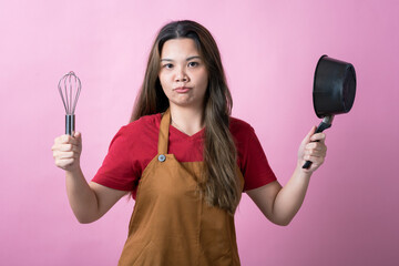 Asian woman in a red shirt and brown apron holds a metal whisk in one hand and a small black pot in the other, smiling confidently against a solid pink background.