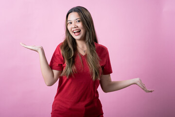 A cheerful young woman with long brown hair and light skin, wearing a red T-shirt, gestures playfully with her palm facing up while smiling and looking downward, against a solid pink background.