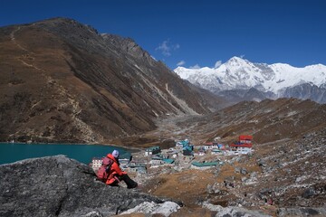 Silhouette of woman in red jacket on viewpoint with amazing panorama of Gokyo with turquoise lake among mountains in background and buildings of settlement. Himalayas, Nepal