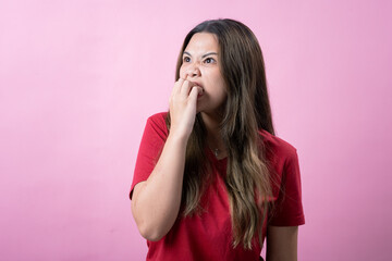 Asian woman in a red t-shirt standing against a pink background, showing a worried or nervous expression while biting her nails and frowning, depicting stress, anxiety