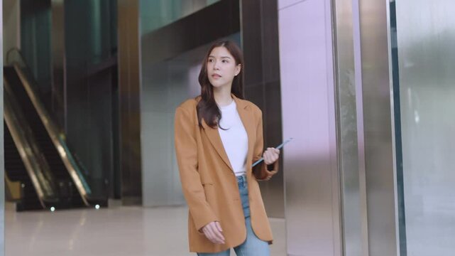 Asian Business Modern business woman in brown blazer with tablet near elevator in glass building. Remote work. Businesswoman walking in modern office. Female Leader and Leadership skills