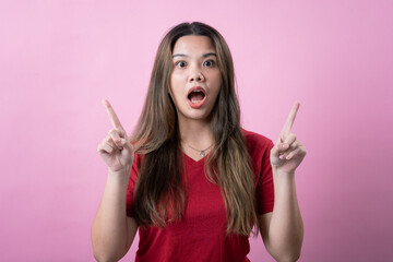 Obraz premium Asian woman in a red t-shirt standing against a pink background, smiling widely with joy, raising both index fingers upward, expressing excitement, happiness