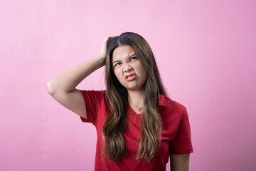 Young woman in a red shirt scratches her head with a confused or frustrated expression against a pink background. Her body language and facial expression convey uncertainty or irritation.