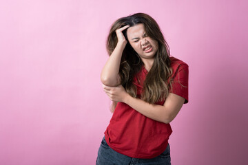 Fototapeta premium Asian woman in a red shirt with long brown hair grimaces in frustration, holding her head with one hand and grabbing her elbow with the other, standing against a solid pink background.