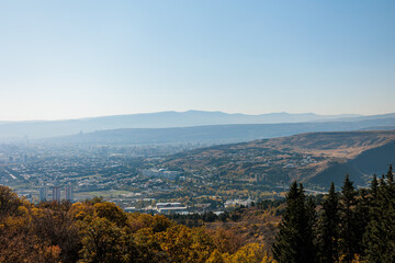 Panoramic autumn view of Tbilisi, Georgia, with its urban landscape stretching towards distant mountains under a clear blue sky. A captivating blend of cityscape and natural scenery