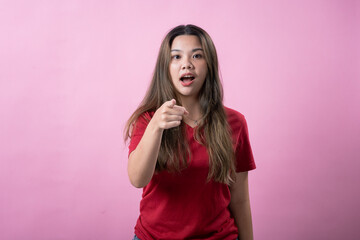 Asian woman with long brown hair and red shirt points directly at the camera with a surprised expression, standing against a solid pink background.