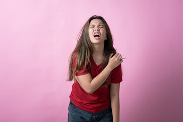 Asian woman in a red shirt with long brown hair grimaces in pain while holding her shoulder, standing against a solid pink background.