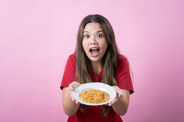 Surprised young woman with long brown hair wearing a red t-shirt, holding a white plate filled with instant noodles, posing against a pink background. Perfect for food related concepts.