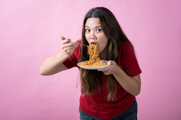 Asian woman in a red shirt with long brown hair eagerly eating noodles from a white plate with a fork, against a solid pink background.