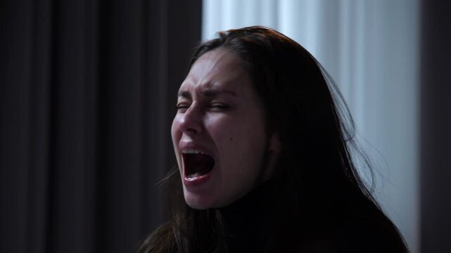 Woman suffering mental health issues, sitting at the table in front of camera, depressed expression, starts crying hysterically laughing, close shot.