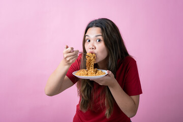 Asian woman in a red t-shirt eating noodles with a fork and holding a plate, her wide eyes and playful expression suggesting excitement or enjoyment