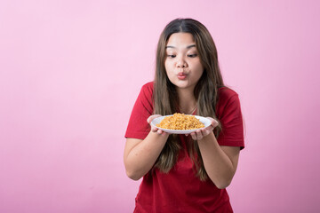 Surprised young woman with long brown hair wearing a red t-shirt, holding a white plate filled with instant noodles, posing against a pink background. Perfect for food related concepts.
