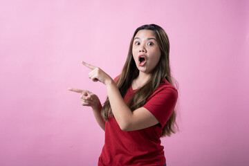 Naklejka premium Asian woman in a red t-shirt standing against a pink background, with wide eyes and mouth open in surprise, pointing dramatically to the side with both hands