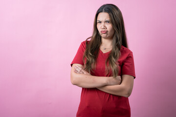 Asian woman in a red shirt with long brown hair crosses her arms and makes a pouty, dissatisfied expression against a vibrant pink background.