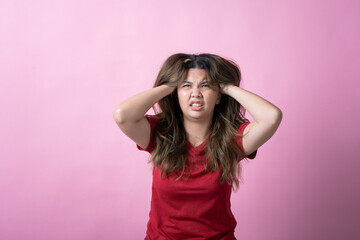 Fototapeta premium Asian woman in a red shirt with long brown hair grips her head with both hands, eyes closed, and mouth open in frustration against a pink background.