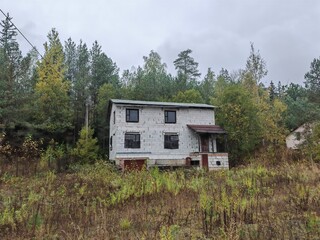 An old unfinished and abandoned silicate block house with a metal roof and boarded up windows is surrounded by a lawn with weeds and woods. There is a wooden awning over the front door. Overcast