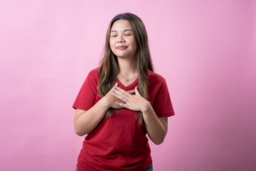 A young woman with long brown hair and light skin, wearing a red T-shirt, places her hands over her chest with a sincere and thoughtful expression, standing against a solid pink background.