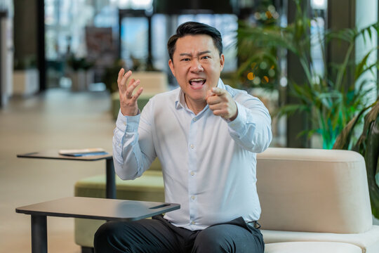 Angry mature boss in shirt arguing over video call looking at camera in modern workplace in office. Unsatisfied Chinese businessman manager communicate with subordinates about the company problems. - Powered by Adobe