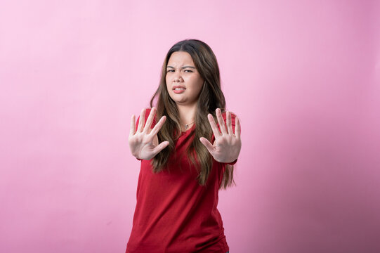 A young woman with long brown hair and light skin, wearing a red T-shirt, makes a disgusted expression while raising her hands defensively, standing against a solid pink background.
