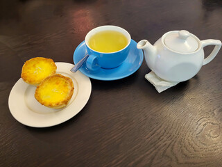 There are two baked baskets on a white plate, a white teapot, a blue cup with green tea on a saucer
