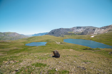 Two lakes, sunlit green meadow, alpine valley and mountain range. Blue sky gradient reflected in water. Top view from rocks to dirt road between two beautiful lakes in high mountains in sunny day.