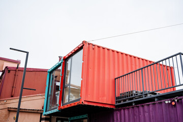 There is an eyecatching red and purple shipping container that features a balcony placed on top,...