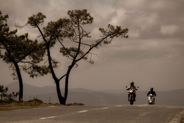 Dos motociclistas disfrutando de un paseo por una carretera rural rodeada de naturaleza, bajo un cielo nublado.