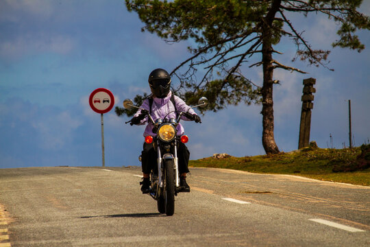 Primer plano de una chica motociclista avanzando por una carretera rural con un cartel de prohibido adelantar al fondo.