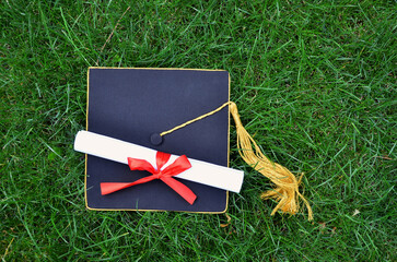 Black master's cap with a golden tassel , graduation hat and diploma on green grass background. Top view outdoors photo. Education ,finishing studying concept. Free copy space.