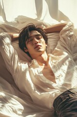 Obraz premium Young asian male relaxing in sunlit bedroom with white shirt.