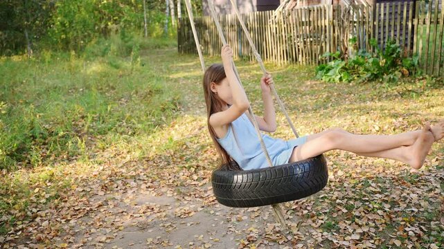 Girl swings barefoot on homemade swing made from rubber car tire, suspended from tree. Happy girl plays outside, enjoys country vacation, swinging with her arms raised up. Games, sports activities.