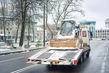 Skid loader transported on truck during winter, construction logistics in cold weather conditions. Loader vehicle on transport truck in snowy city, industrial logistics. Tow truck transporting loader