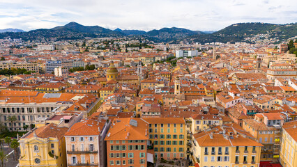 A breathtaking aerial view of Nice, France, showcasing its iconic terracotta rooftops, colorful architecture, and a scenic backdrop of lush green hills and mountains under a partly cloudy sky